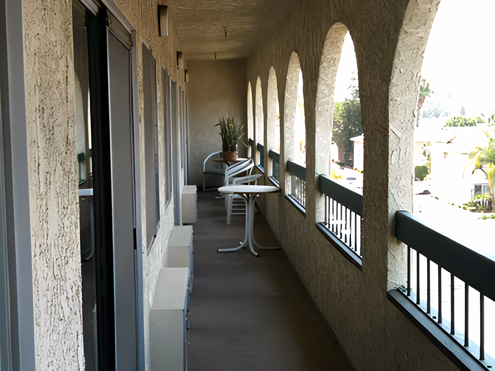A long outdoor balcony with arched openings and a railing overlooking a residential area. The balcony has a small round table, several white chairs, and a potted plant at the far end. The walls and ceiling have a textured finish.