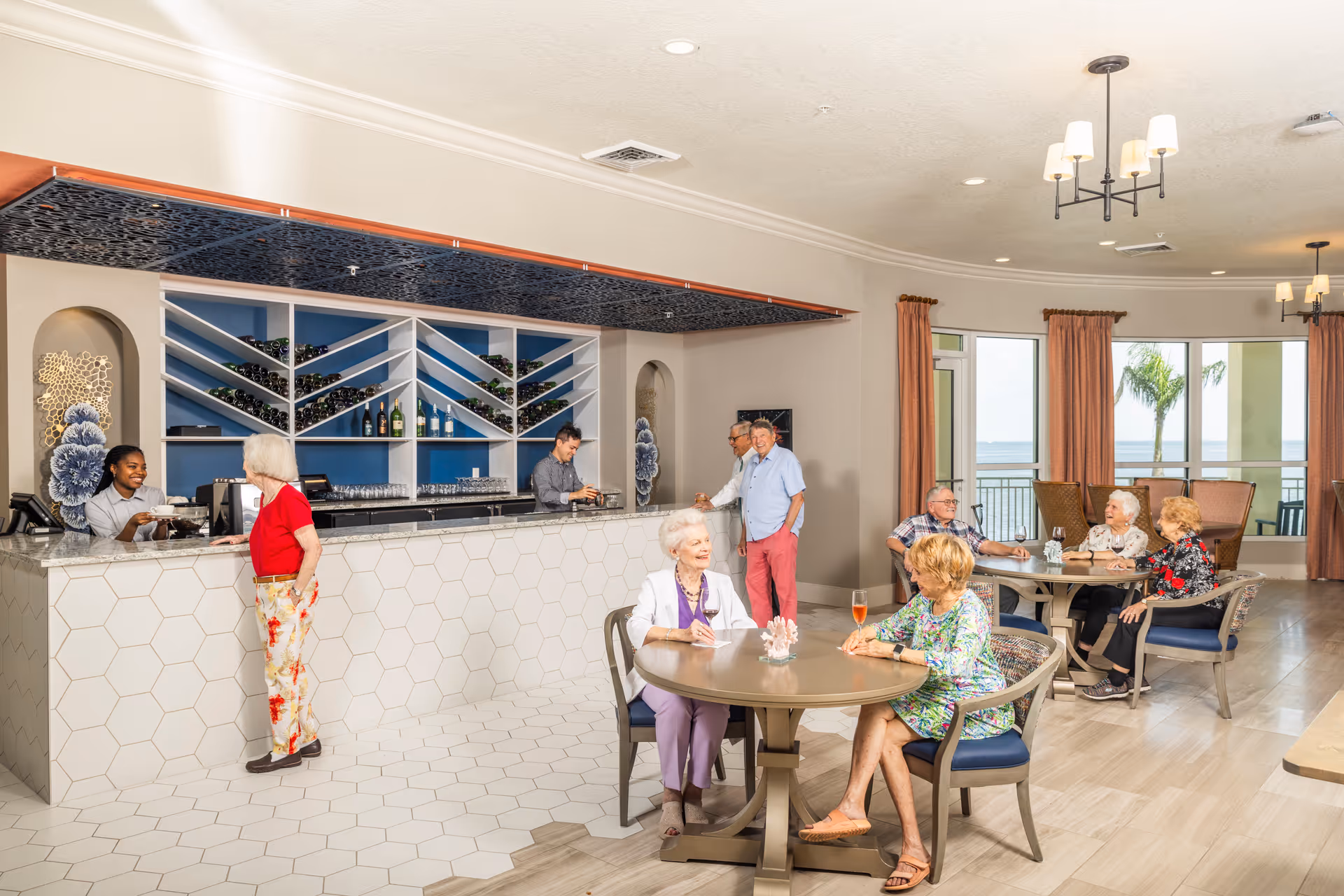 A bright and spacious dining area in a senior living facility with a modern bar counter on the left where two staff members serve an elderly woman. Several elderly residents are seated at round tables, engaging in conversation and enjoying drinks. Large windows in the background offer a view of palm trees and water outside. The room features light-colored walls, hexagonal tile flooring near the bar, and warm lighting fixtures.
