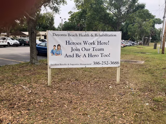 Outdoor view of a sign for Daytona Beach Health & Rehabilitation Center placed on a grassy area with trees and parked cars in the background. The sign reads: 'Heroes Work Here! Join Our Team And Be A Hero Too! Excellent Benefits & Supportive Management 386-252-3686' and includes an image of two healthcare workers.