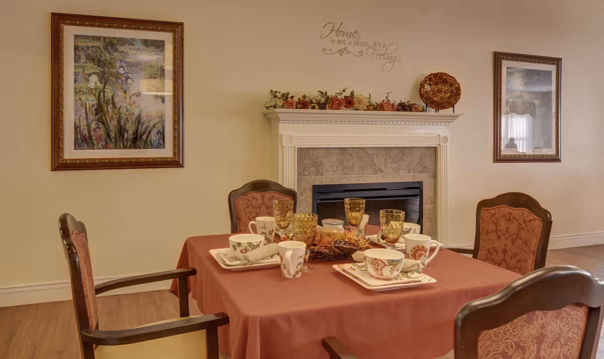 A cozy dining area with a table set for tea in front of a fireplace and framed artwork on the walls.