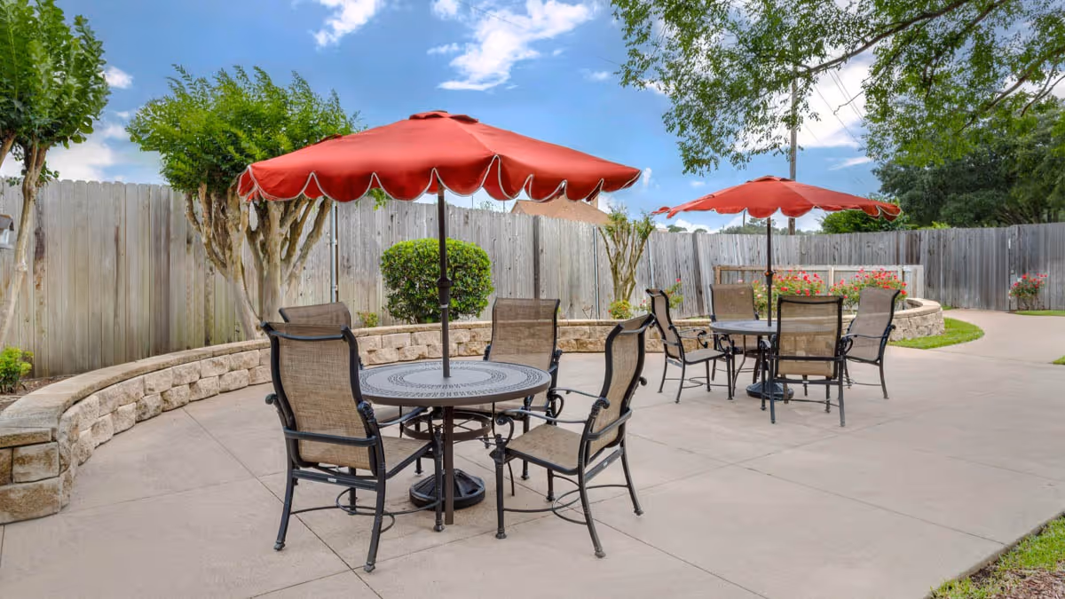 Outdoor patio with round metal tables and chairs under red umbrellas, a low stone wall, and a wooden fence.