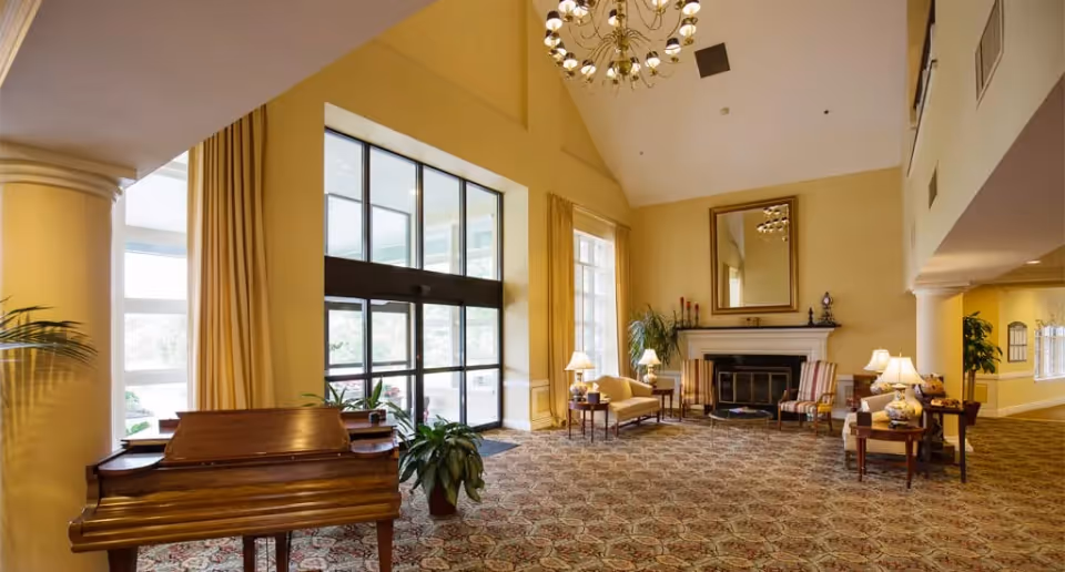 Spacious senior living lobby with high ceilings, a chandelier, fireplace, piano, and seating near large glass entrance doors.