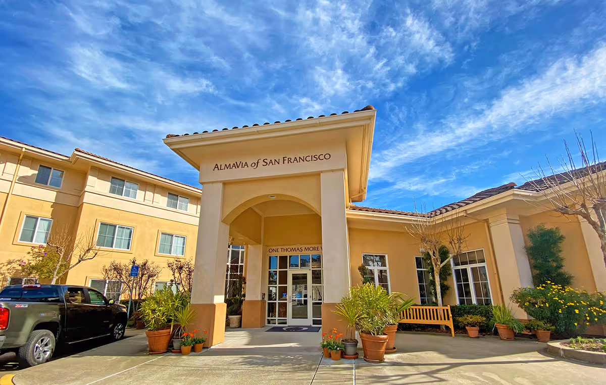 Exterior front entrance of AlmaVia of San Francisco facility with a covered walkway, potted plants, a bench, and a clear blue sky with scattered clouds above.