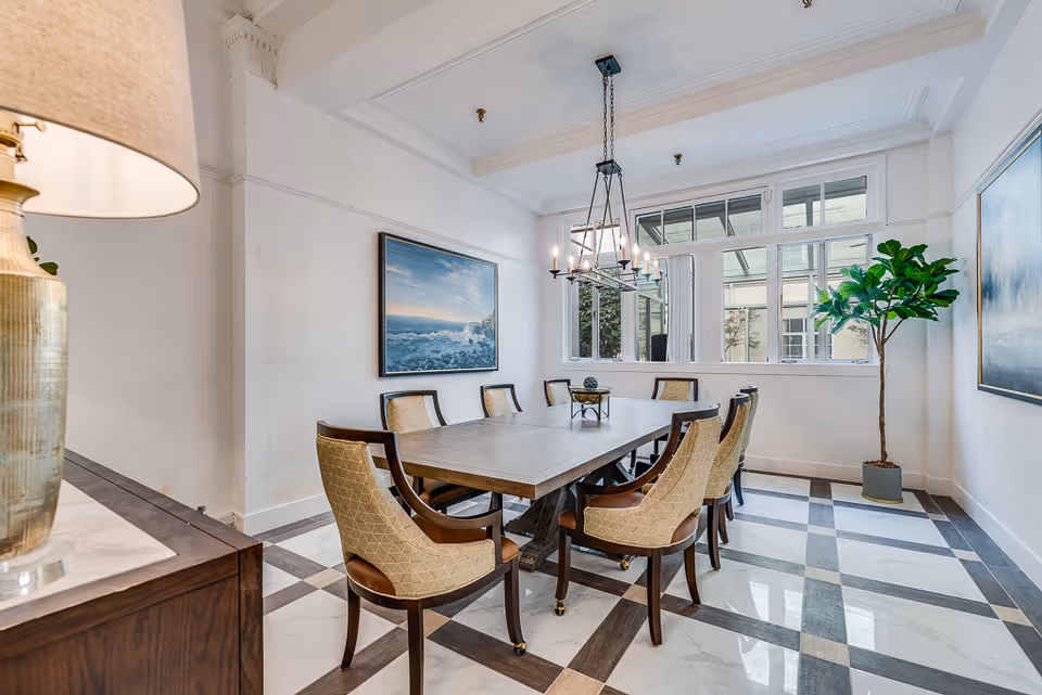 A bright dining room with a large rectangular wooden table surrounded by eight upholstered chairs. The room features a checkered tile floor, white walls, a large window letting in natural light, a modern chandelier hanging above the table, a potted plant in the corner, and framed artwork on the walls.