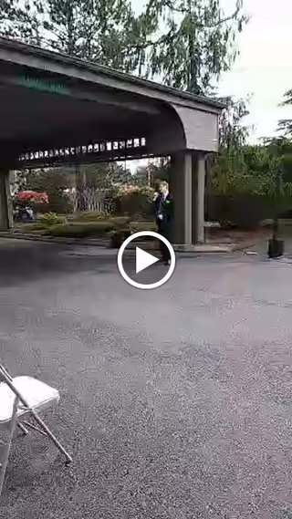 Covered porte-cochere and driveway at a senior living entrance with a person near the columns and a folding chair in the foreground.