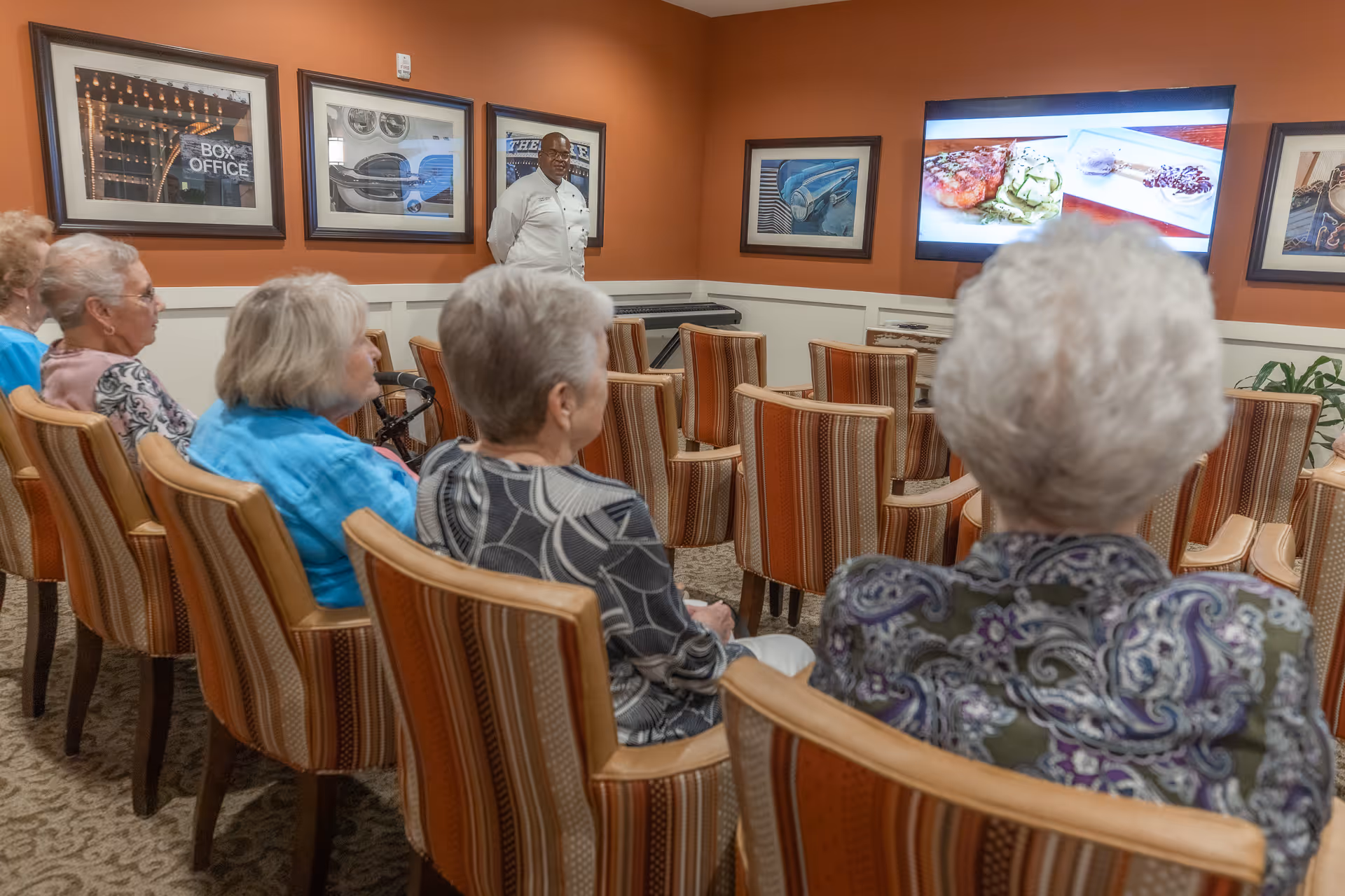 A group of elderly women seated in striped chairs facing a television screen in a room with orange walls. A man in a white chef's coat stands near the front of the room. The television screen displays images of food. Framed pictures hang on the walls.