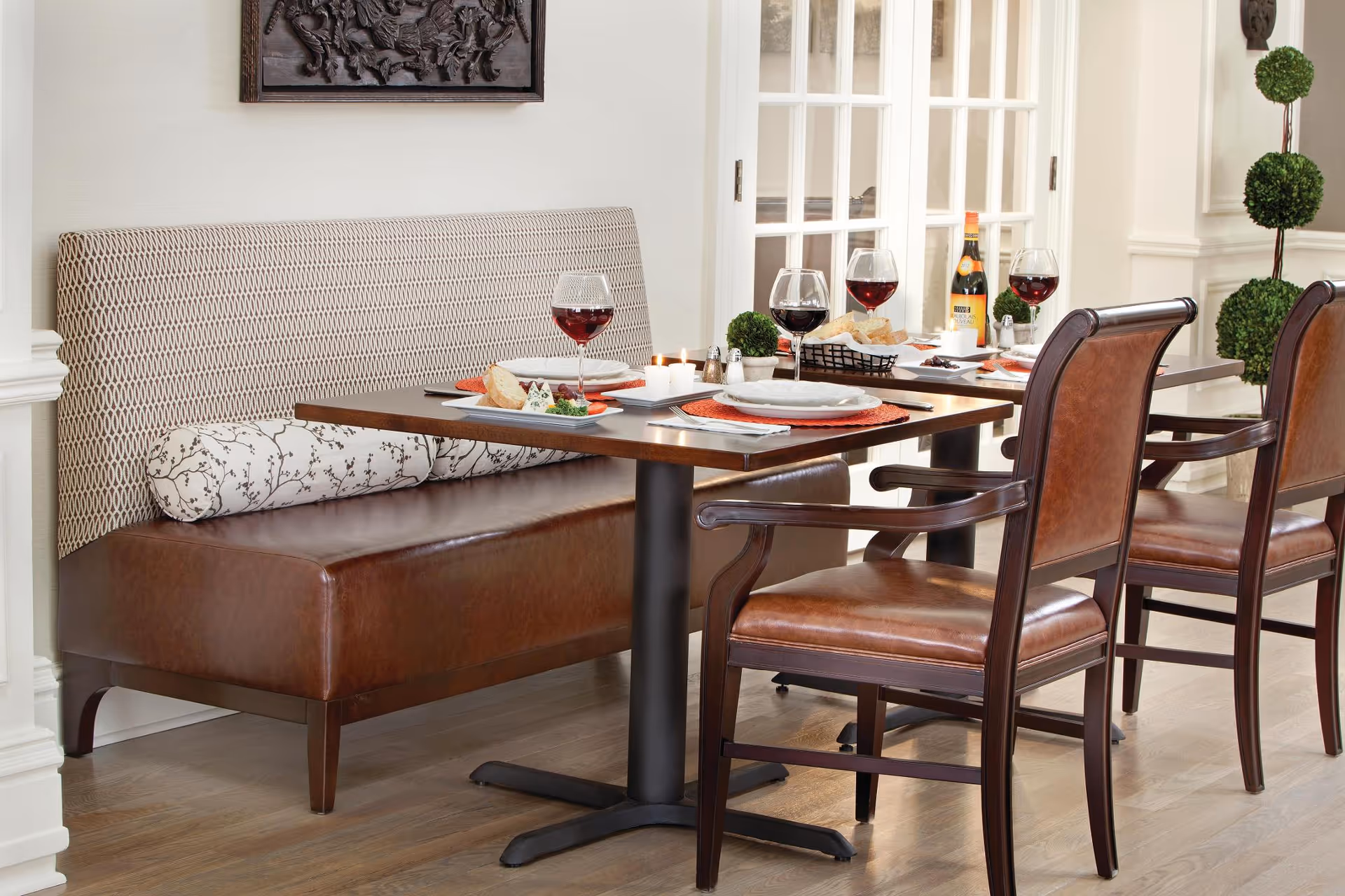 A dining area with a wooden table set for four, featuring a cushioned bench with patterned upholstery on one side and three wooden chairs with leather seats on the other. The table is set with plates, wine glasses filled with red wine, a bottle of wine, bread, and small decorative plants. The background includes white walls, a framed artwork, and a decorative topiary.