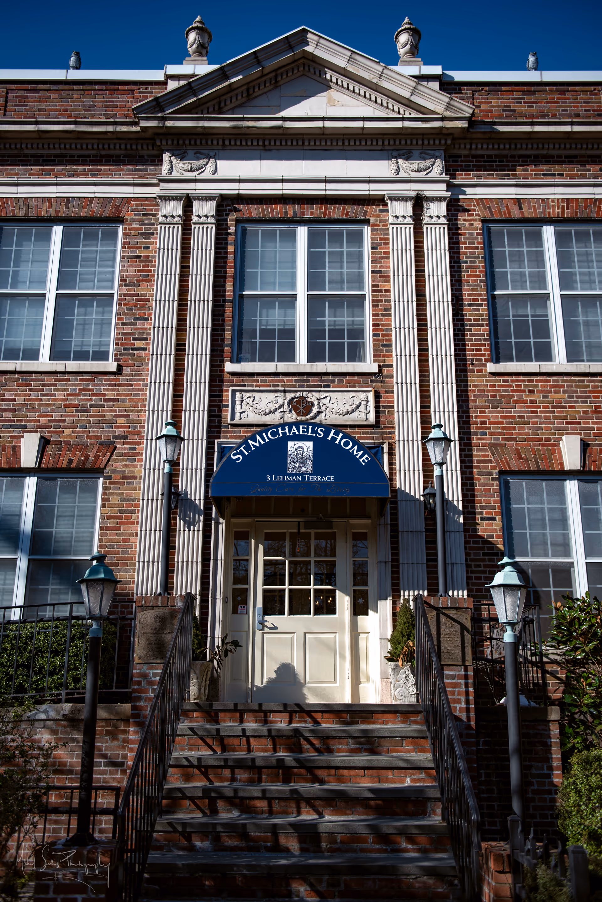 Front entrance of St Michael's Home, a brick building with white trim and columns. There are steps leading up to a white door with a blue awning above it displaying the facility name and address. Four vintage-style lamp posts flank the stairs and entrance.