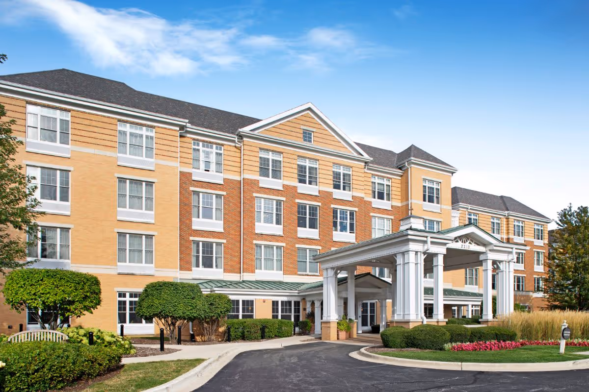 Front entrance of a multi-story senior living building with a columned porte-cochere, driveway, and landscaped shrubs and flowers.