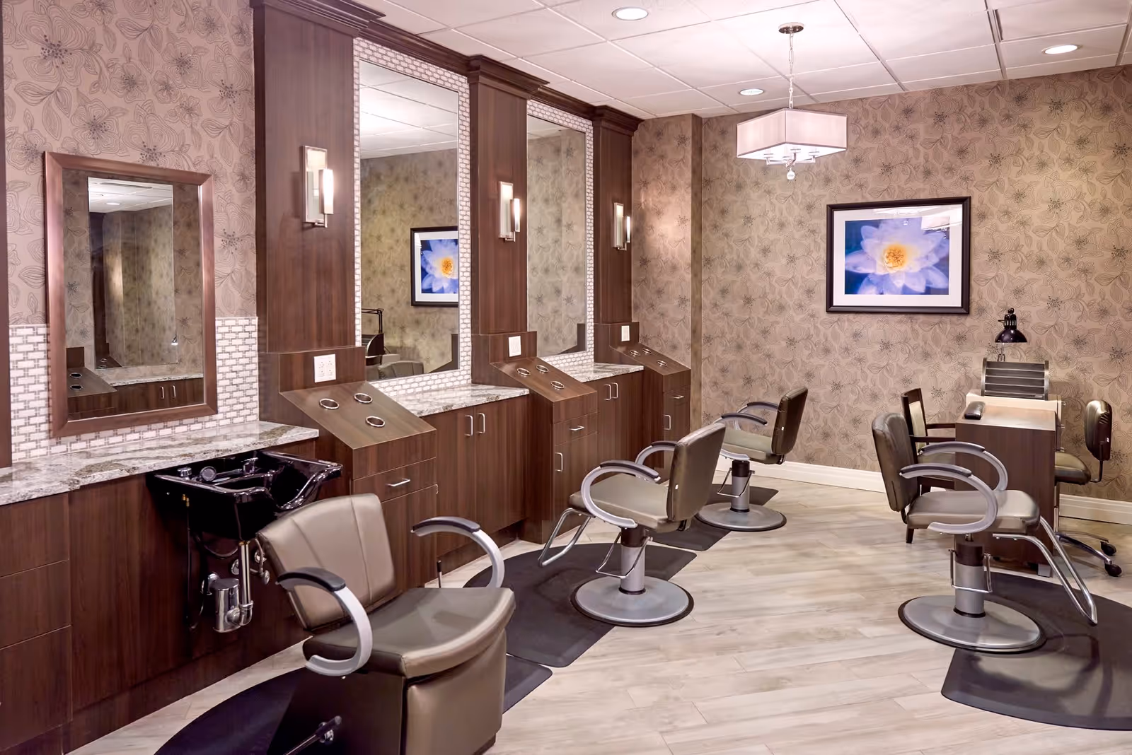 Interior view of a salon area in an assisted living facility featuring multiple styling chairs in front of large mirrors mounted on wooden cabinetry. The room has floral patterned wallpaper, a framed flower picture on the wall, and a manicure station with chairs and a desk lamp. The floor is light-colored wood, and the ceiling has recessed lighting and a hanging light fixture.