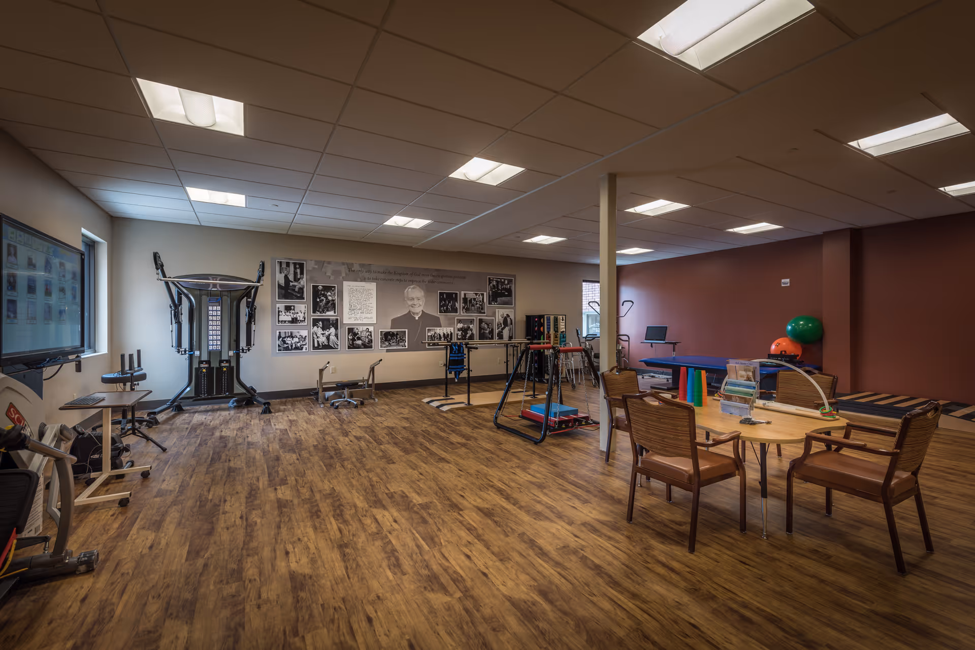 A spacious room with wooden flooring and a drop ceiling with fluorescent lights. The room contains various exercise and therapy equipment including a multi-gym machine, parallel bars, therapy balls, and a therapy table. There is a round table with four chairs and some colorful cones on it. The walls have framed black and white photos and a large portrait of a man. Two windows allow natural light into the room.