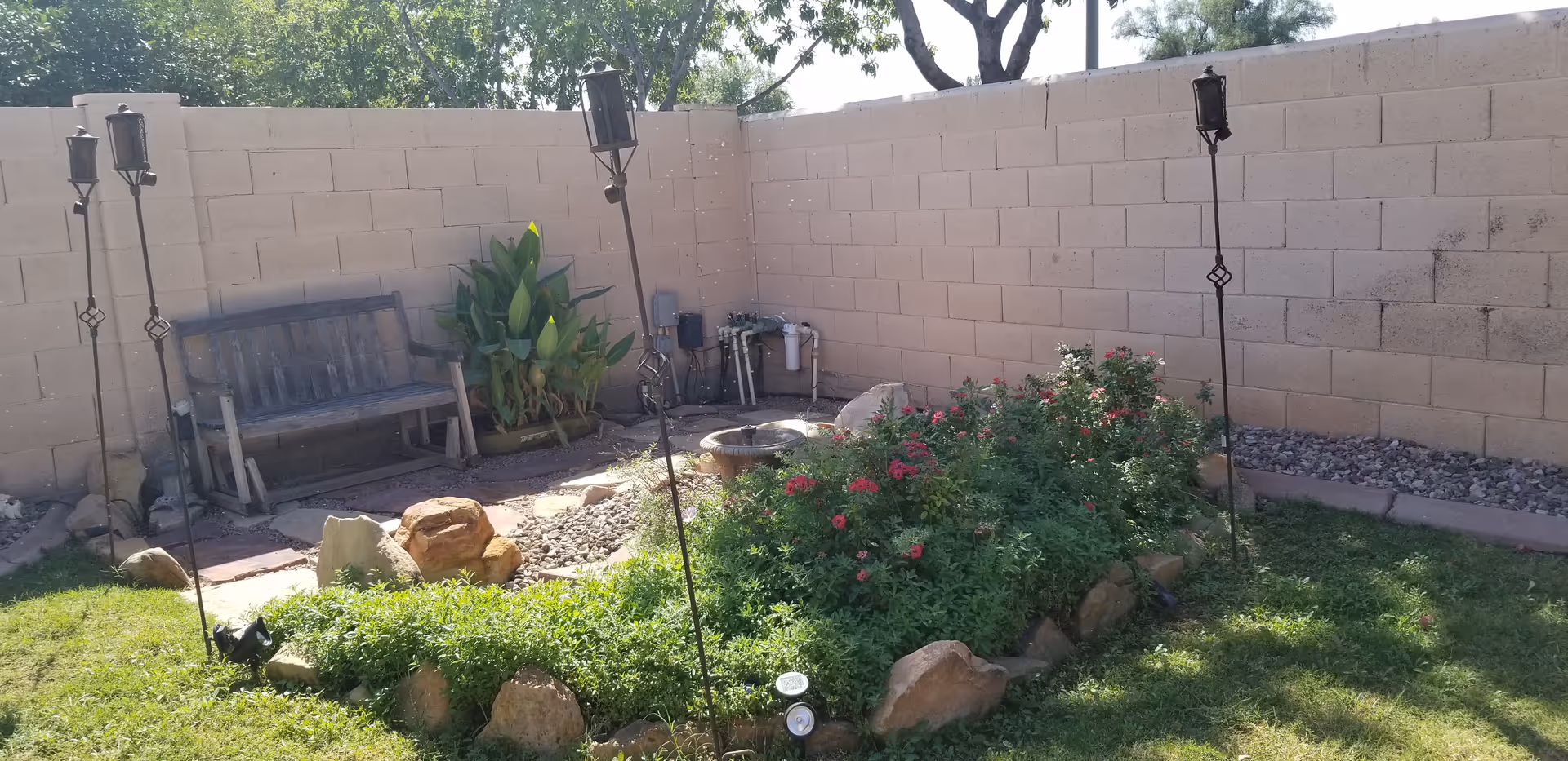 A small outdoor garden area enclosed by a beige brick wall, featuring a wooden bench, several tall plants, a flower bed with red flowers surrounded by rocks, four tall tiki torches, and a small water fountain in the corner.