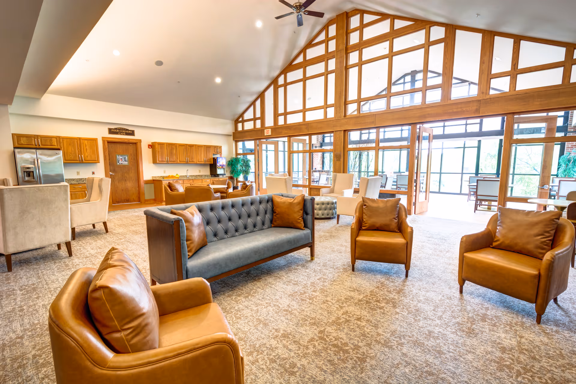A spacious and well-lit living room area in a senior living facility with a high vaulted ceiling and large windows. The room features a gray tufted sofa with brown pillows, three brown leather armchairs, a carpeted floor, and a ceiling fan. In the background, there is a kitchenette with wooden cabinets, a refrigerator, and a dining area with tables and chairs near large glass doors leading to an outdoor patio.
