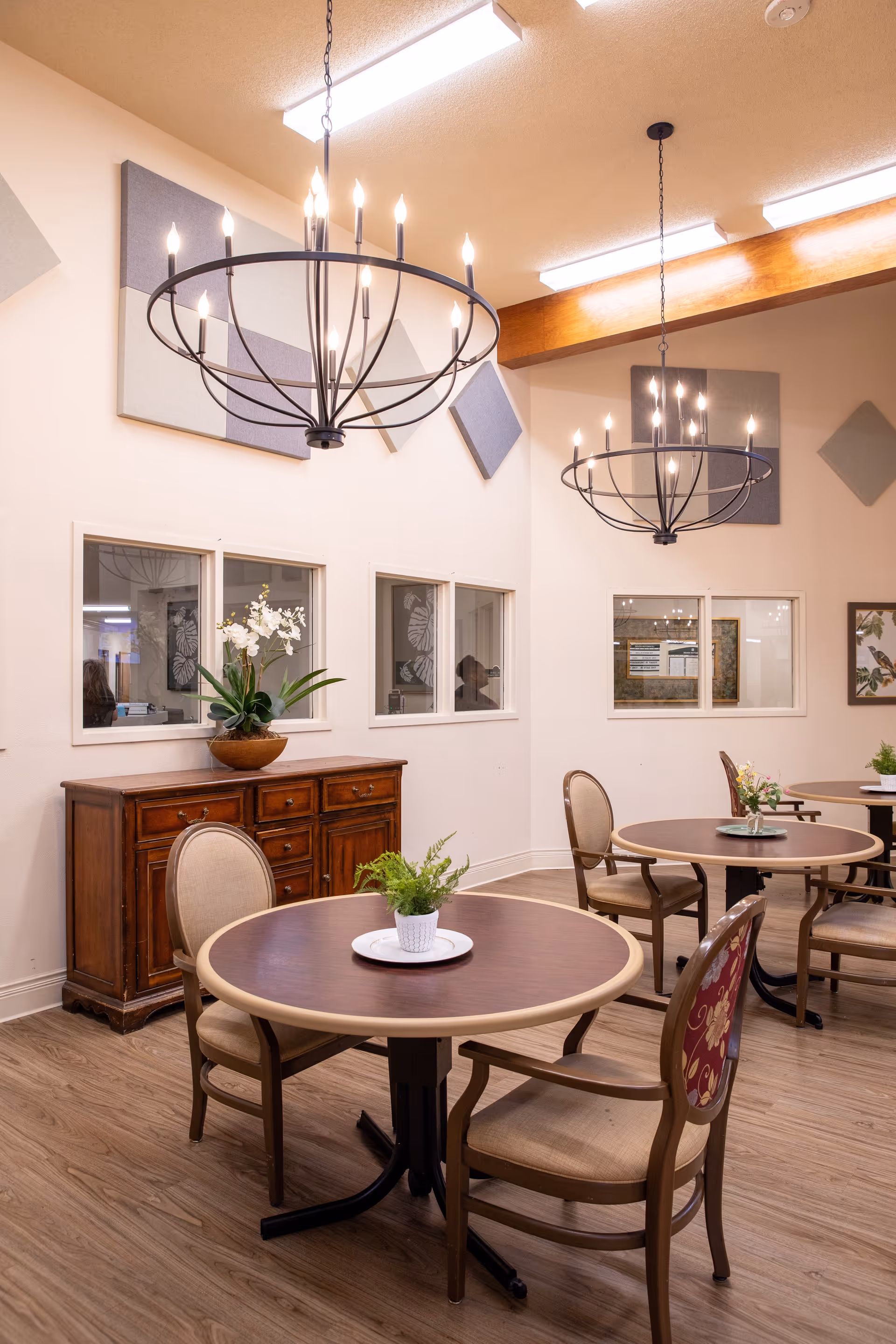 A dining area with round wooden tables and cushioned chairs. Each table has a small plant centerpiece. The room features wooden flooring, a wooden sideboard with a potted orchid, and modern chandeliers hanging from the ceiling. The walls have decorative acoustic panels and windows looking into another room.