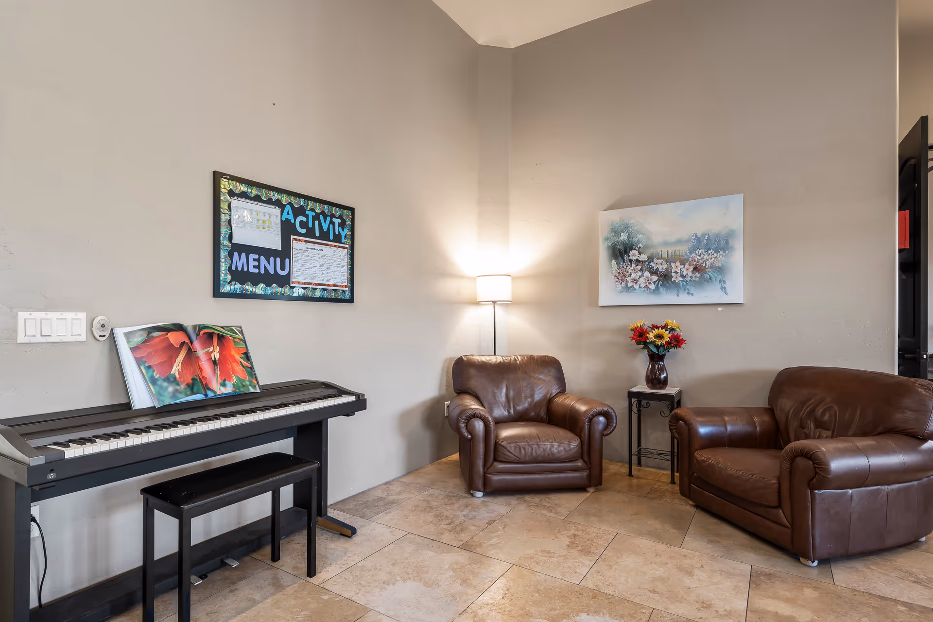 A cozy sitting area in a senior living facility with two brown leather armchairs, a small side table with a vase of colorful flowers, a floor lamp, a digital piano with a bench, and a bulletin board labeled 'Activity Menu' on the wall.
