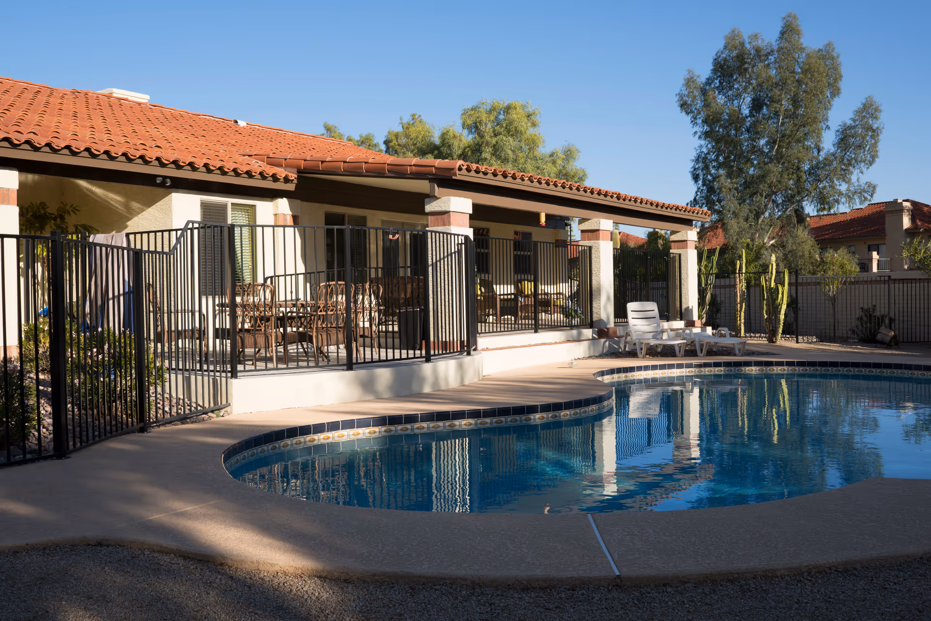 Outdoor swimming pool area at Blue Lakes Assisted Living with a curved pool surrounded by a concrete deck. There is a black metal fence enclosing a covered patio with outdoor seating and tables. The building has a red tile roof and beige walls, with trees and plants in the background under a clear blue sky.