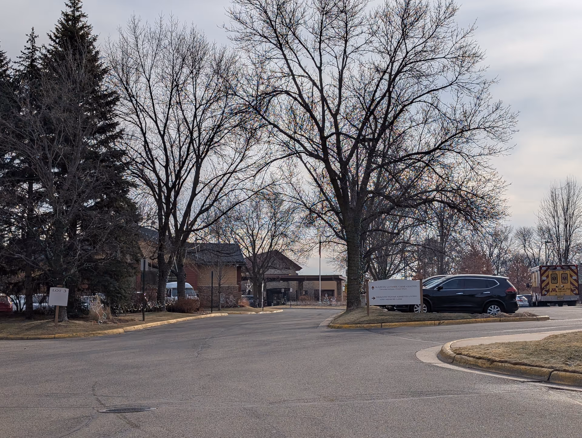 View of the Martin Luther Campus parking lot with several parked vehicles, leafless trees, and a building entrance in the background under a cloudy sky.
