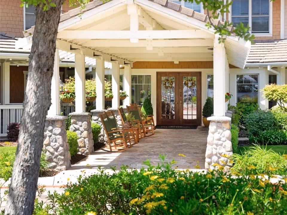 Covered porch entrance of a senior living facility with wooden rocking chairs lined up along stone pillars, surrounded by green plants and flowers, leading to double wooden doors with glass panels and decorative wreaths.
