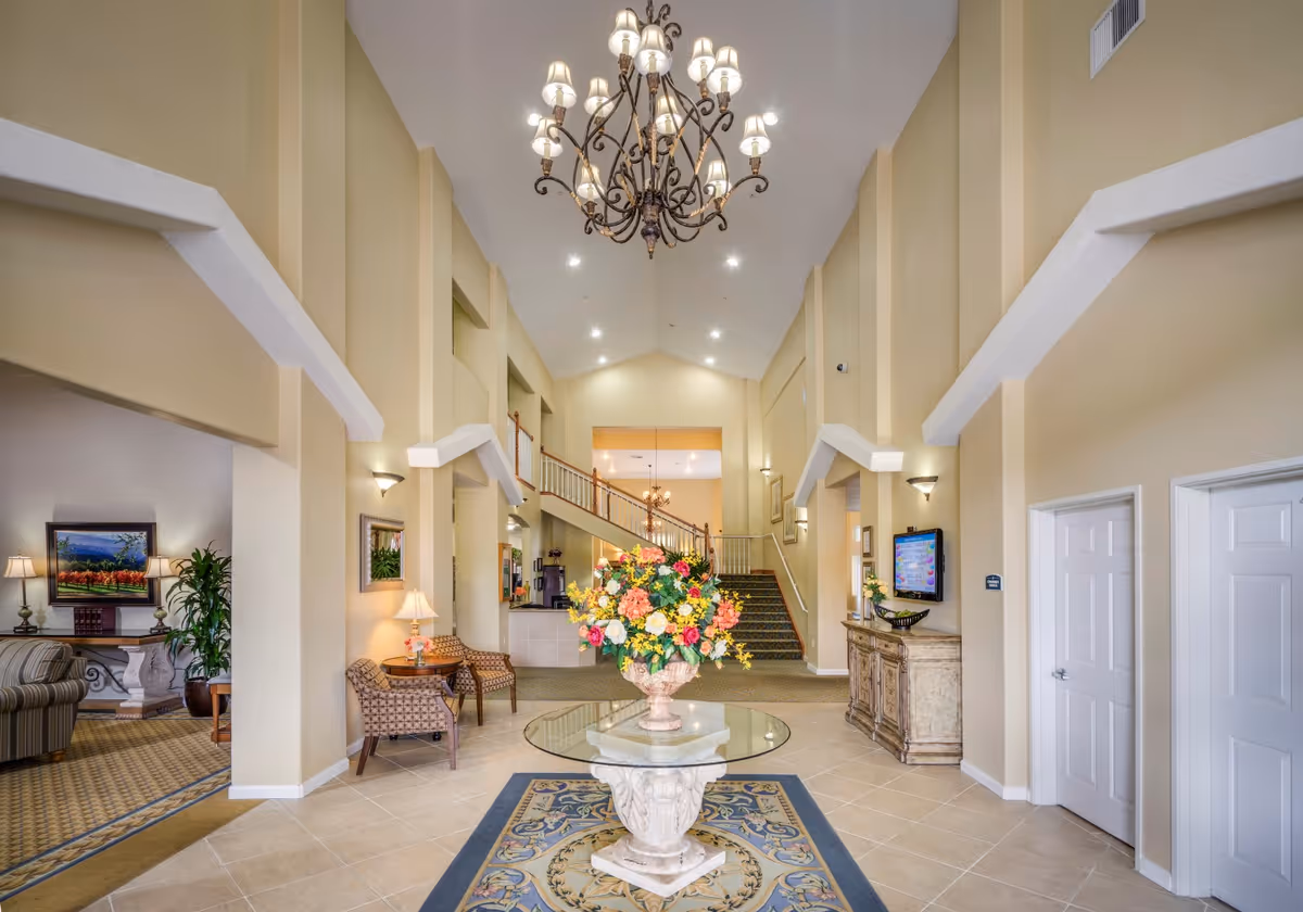 Interior view of a senior living facility lobby with high ceilings, a large chandelier, a round glass table with a floral arrangement in the center, seating areas with chairs and lamps, a staircase leading to an upper level, and neutral-colored walls.