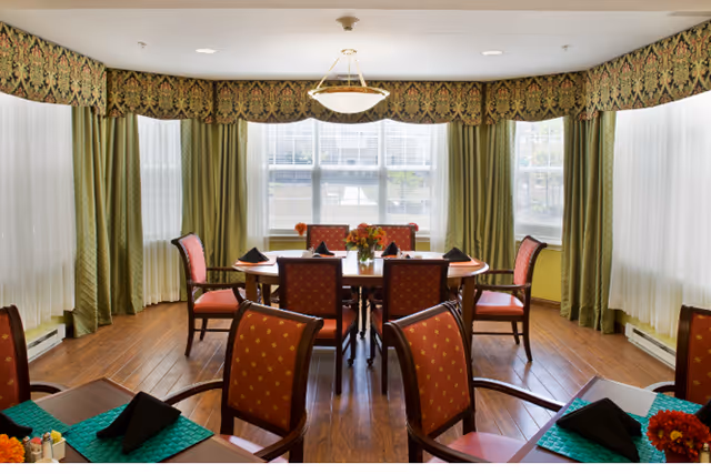 A dining room with a round table surrounded by chairs, decorated with green and red accents. Large windows allow natural light to fill the space, complemented by elegant curtains and a floral centerpiece.