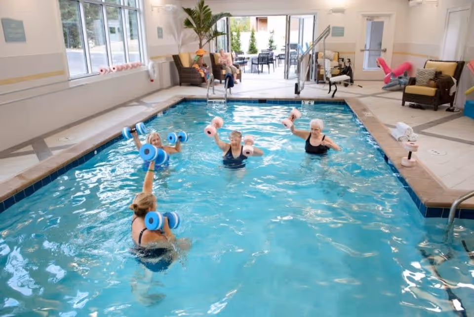 Four elderly women exercising with foam dumbbells in an indoor swimming pool at a senior living facility. The pool area has large windows, chairs, and an open door leading to an outdoor patio with tables and chairs.