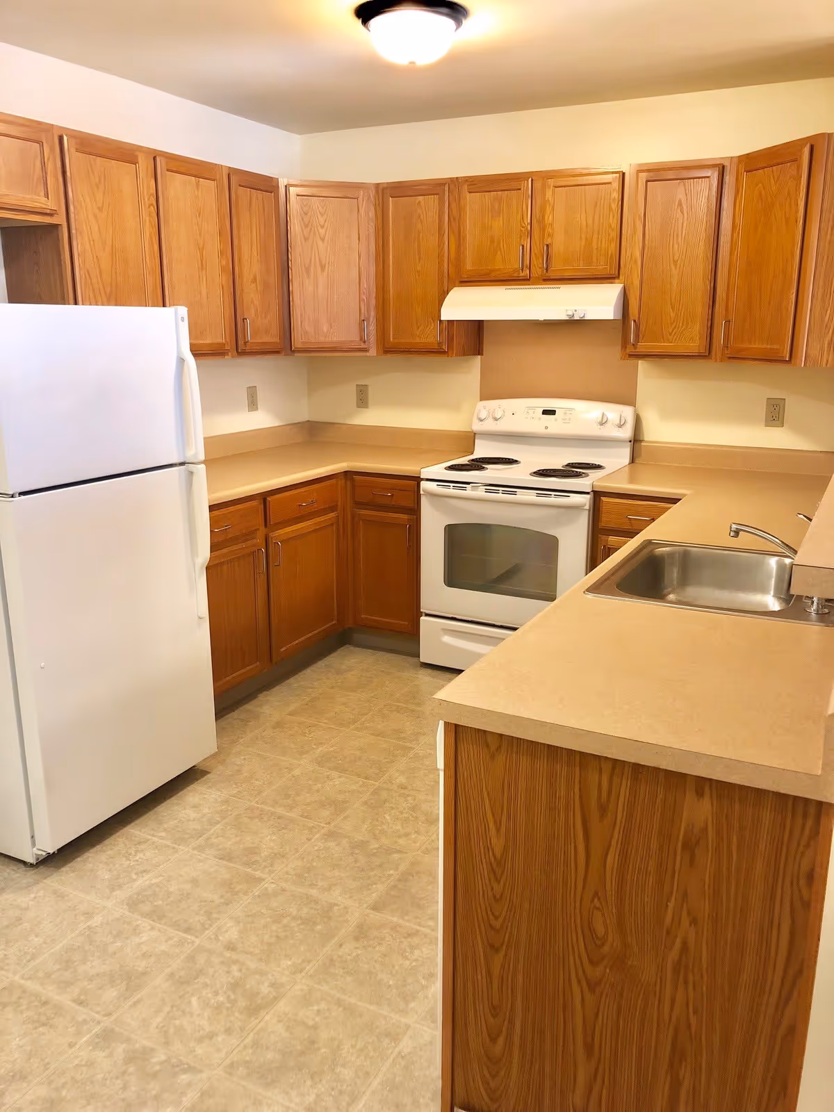 Galley-style kitchen with oak cabinets, beige laminate countertops, a white refrigerator and stove, and a stainless steel sink.