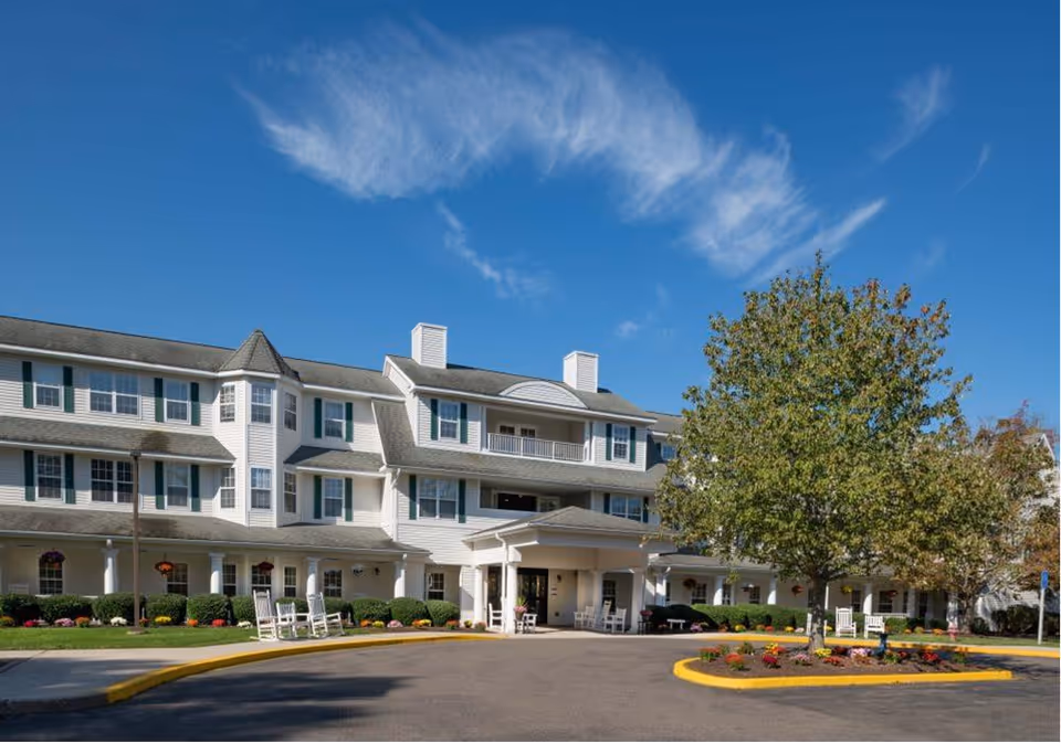 Exterior view of a large, multi-story senior living facility building with white siding and green shutters under a clear blue sky. The entrance has a covered drop-off area with rocking chairs on the porch. There is a landscaped roundabout with a tree and flowers in front of the building.