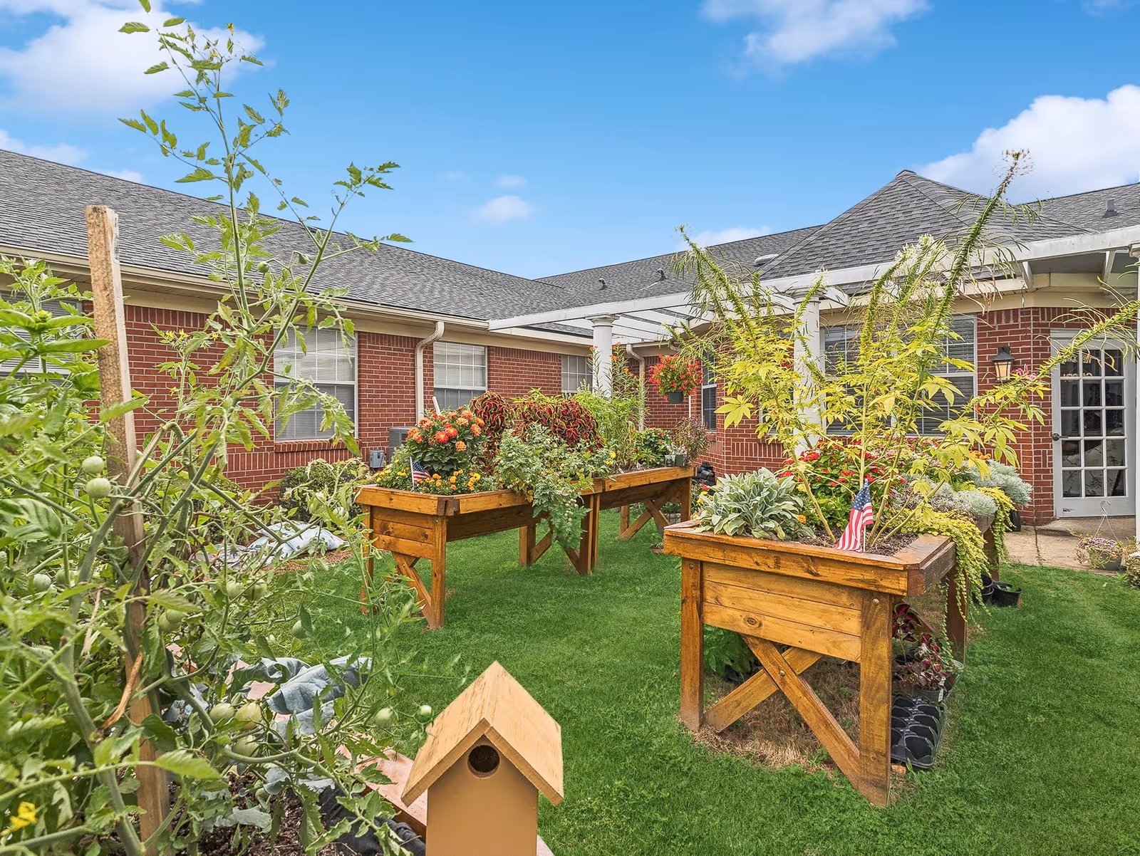 Outdoor garden area at Summer Village Senior Living with raised wooden planter boxes filled with various plants and flowers, green grass, and a small birdhouse in the foreground. The background shows a brick building with windows and a door under a blue sky with some clouds.