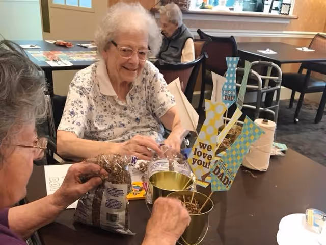 Two elderly women sitting at a table engaged in a craft activity involving small buckets, sticks, and decorative paper cutouts with messages like 'we love you!' and 'DAD'. Another elderly woman is seated in the background near a walker. The setting appears to be a common area in a senior living facility.