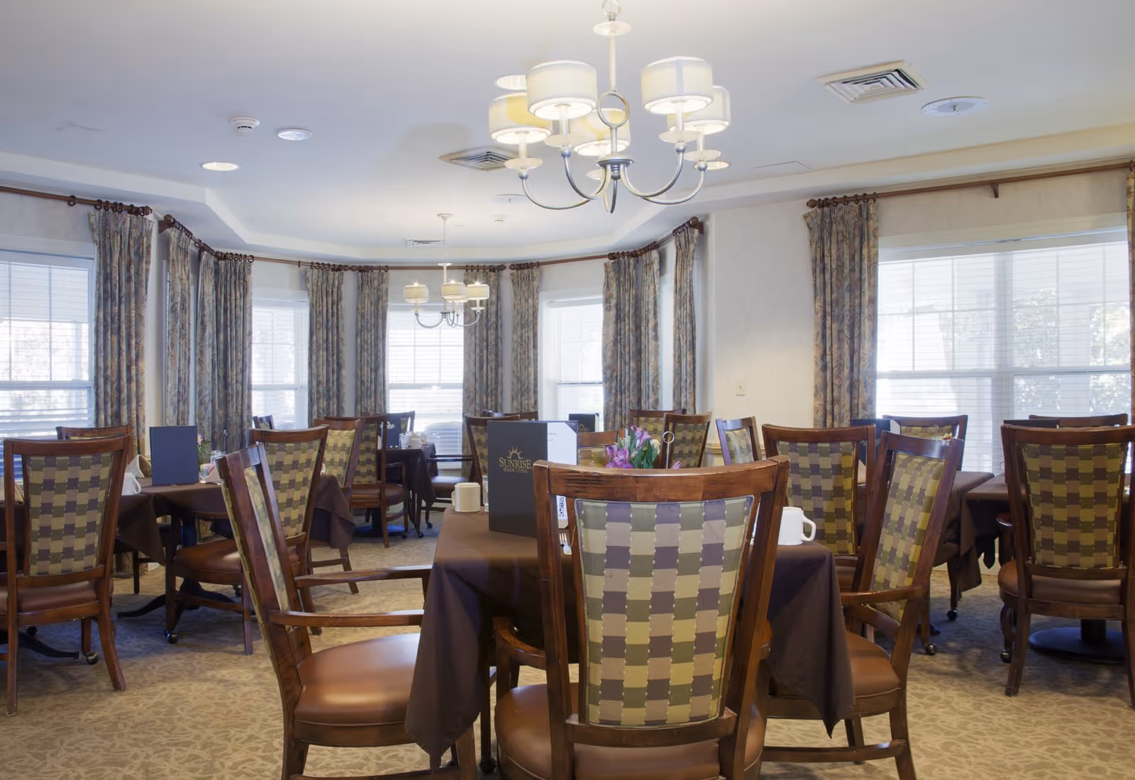 Bright dining room with multiple tables and patterned wooden chairs, large windows with curtains, and chandeliers overhead.
