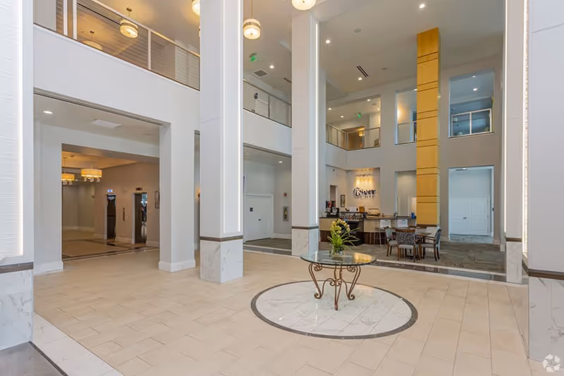 Bright two-story residential lobby with tall columns, a central glass-top table, seating area and a reception desk.