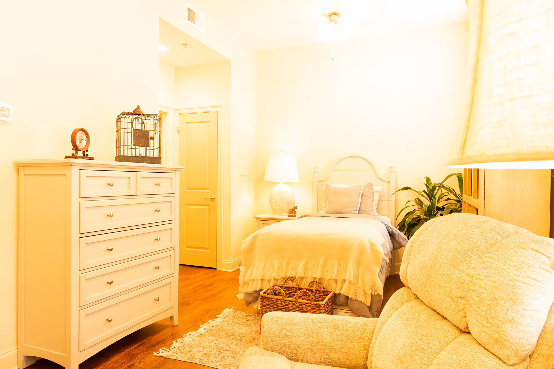 A cozy bedroom in a senior living facility featuring a single bed with beige and gray bedding, a bedside table with a lamp, a white chest of drawers with decorative items on top, a beige armchair, a small rug on wooden flooring, and a potted plant in the corner.