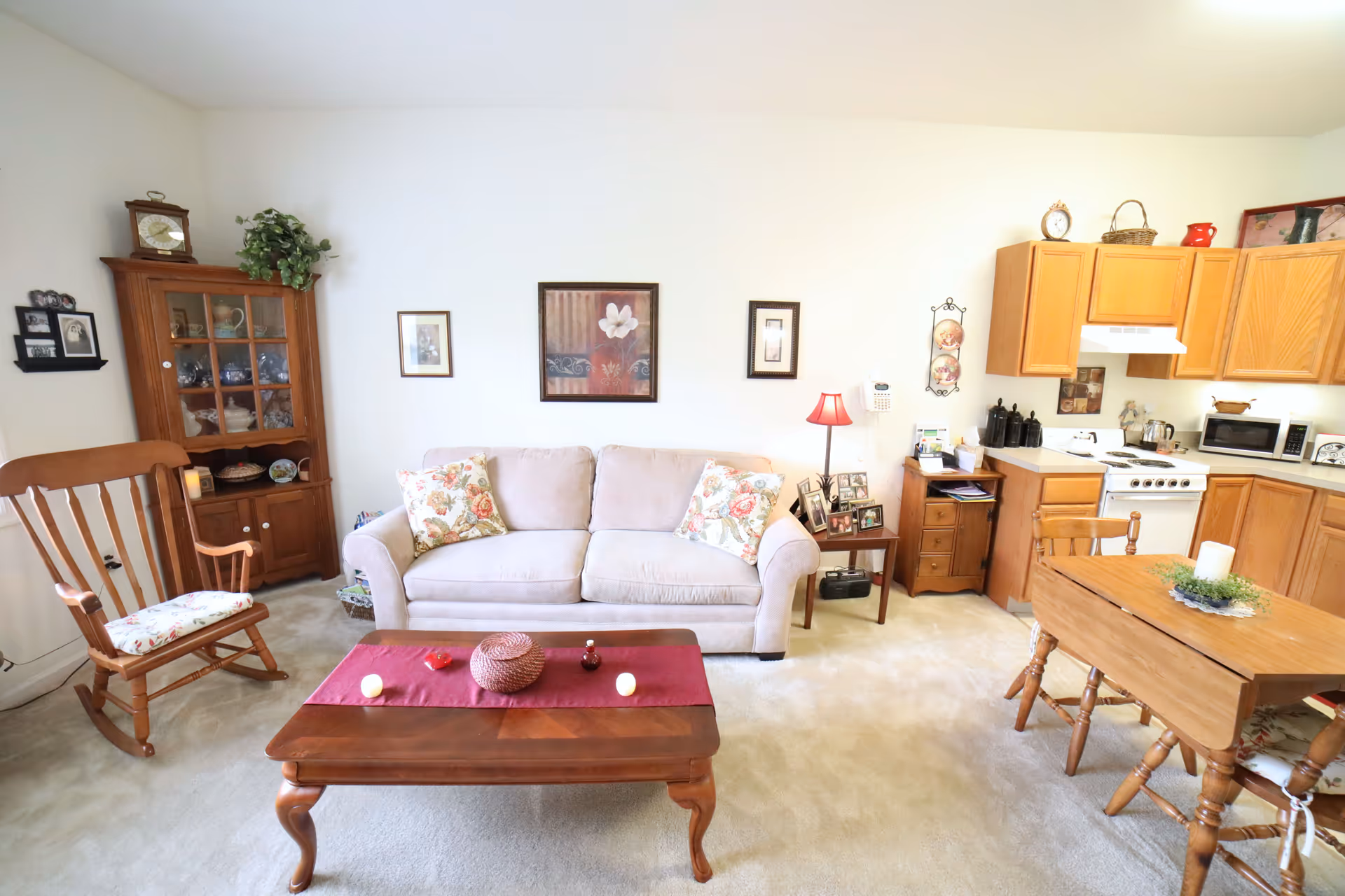 A cozy living room and kitchen area featuring a beige sofa with floral cushions, a wooden rocking chair, a wooden coffee table with decorative items, a wooden dining table with chairs, and a kitchen with wooden cabinets, a stove, microwave, and various kitchen items. The walls are decorated with framed pictures and a clock, and there is a small side table with a red lamp and photo frames.