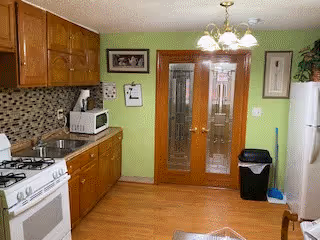A kitchen with wooden cabinets, a white gas stove, a microwave on the countertop, a refrigerator, a trash bin, and double glass doors with wooden frames. The walls are painted light green and there is a decorative tile backsplash behind the stove and countertop.