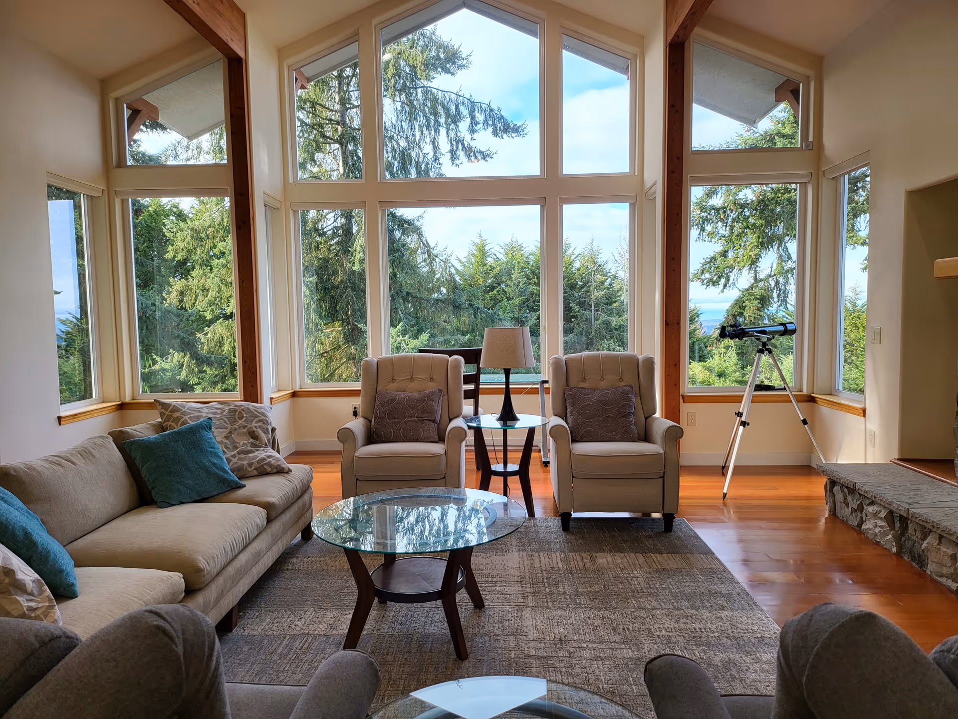 A bright living room with large floor-to-ceiling windows showing a view of tall green trees outside. The room has a beige sofa with blue and patterned pillows, two beige armchairs with patterned cushions, a glass coffee table, a side table with a lamp, a telescope near the window, and a stone fireplace on the right side.