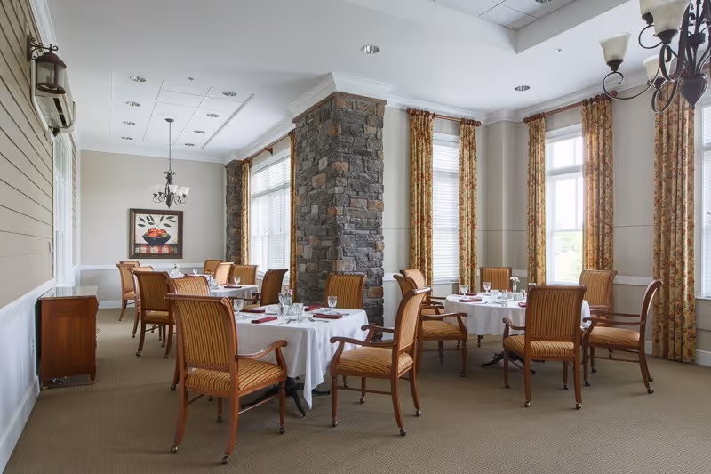 Bright dining room with round tables set with white tablecloths, wooden chairs, tall windows with curtains, and a central stone column.