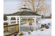 A white wooden gazebo with a shingled roof situated outdoors in a snowy landscape. There is a wooden railing and a small ramp leading up to the gazebo. Leafless trees and snow-covered ground are visible in the background.