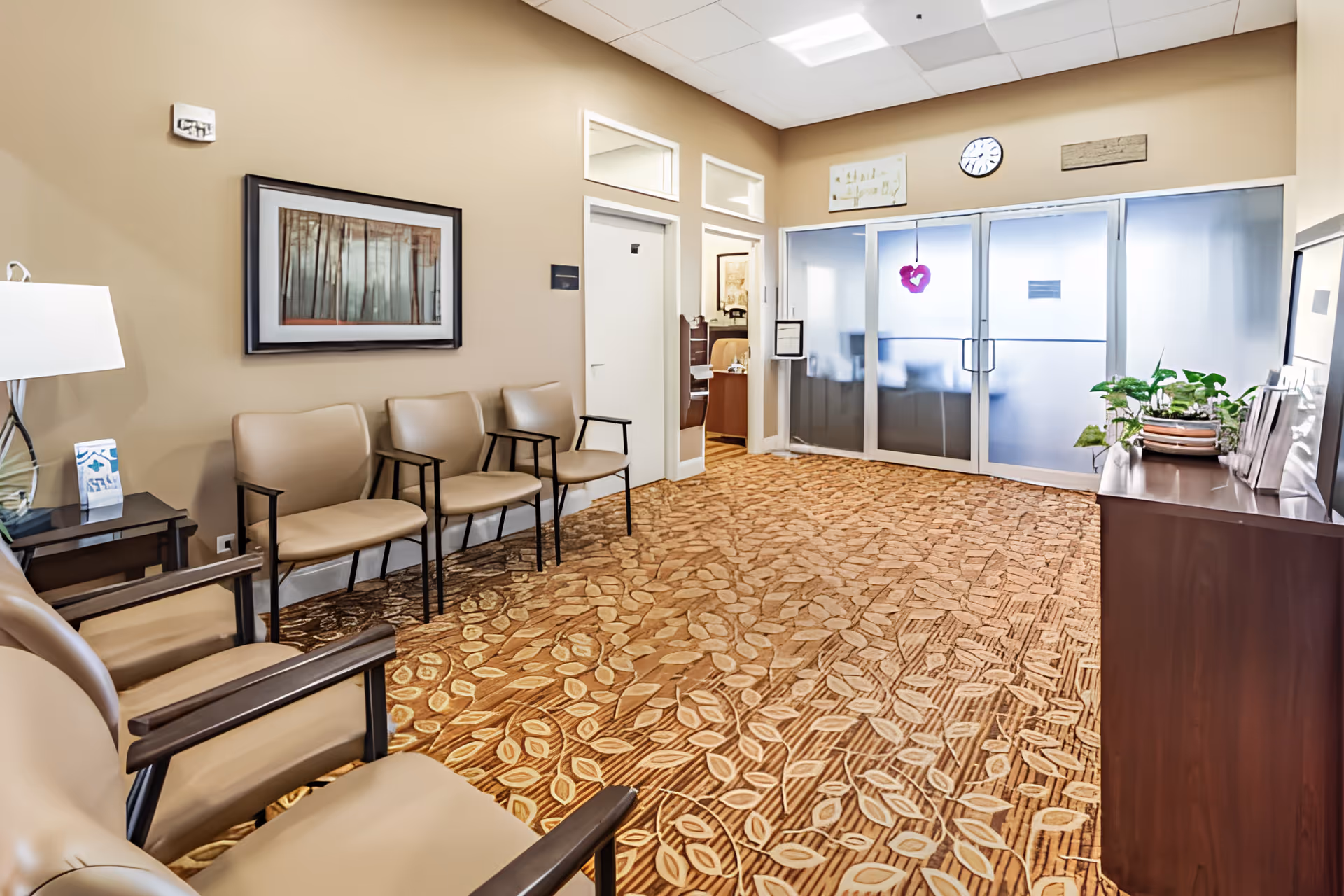 A clean waiting-room interior with beige chairs along the wall, framed artwork, patterned carpet, and frosted glass double doors.