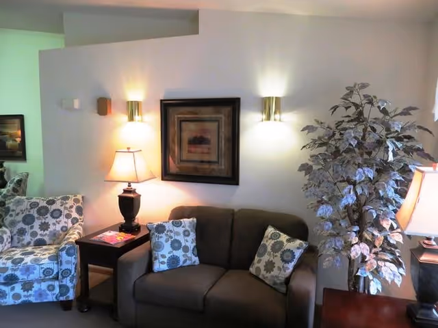 A cozy living room area with a brown loveseat adorned with two floral patterned pillows, a matching floral patterned armchair, a wooden side table with a lamp and magazines, a framed artwork on the wall, two wall-mounted lights, and a large potted plant in the corner.