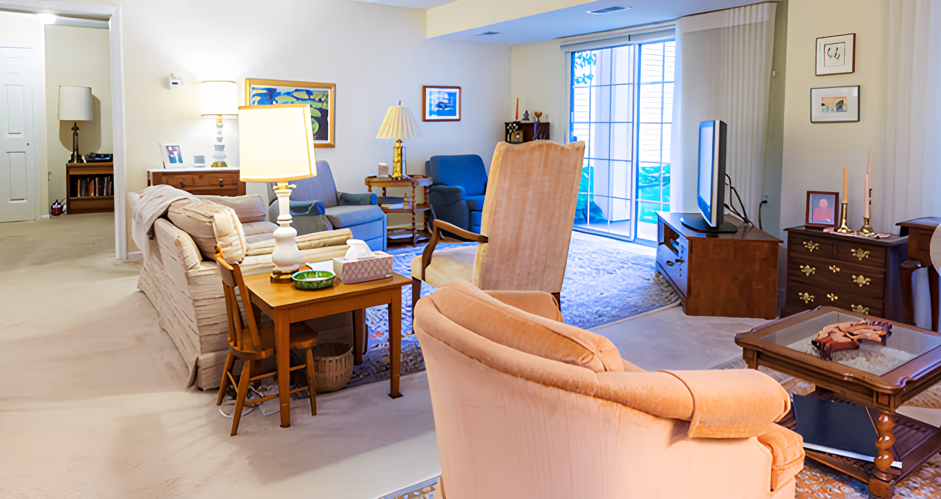 Bright furnished living room with upholstered chairs and sofa, side tables and lamps, a TV on a wooden stand, and a sliding glass door to a patio.