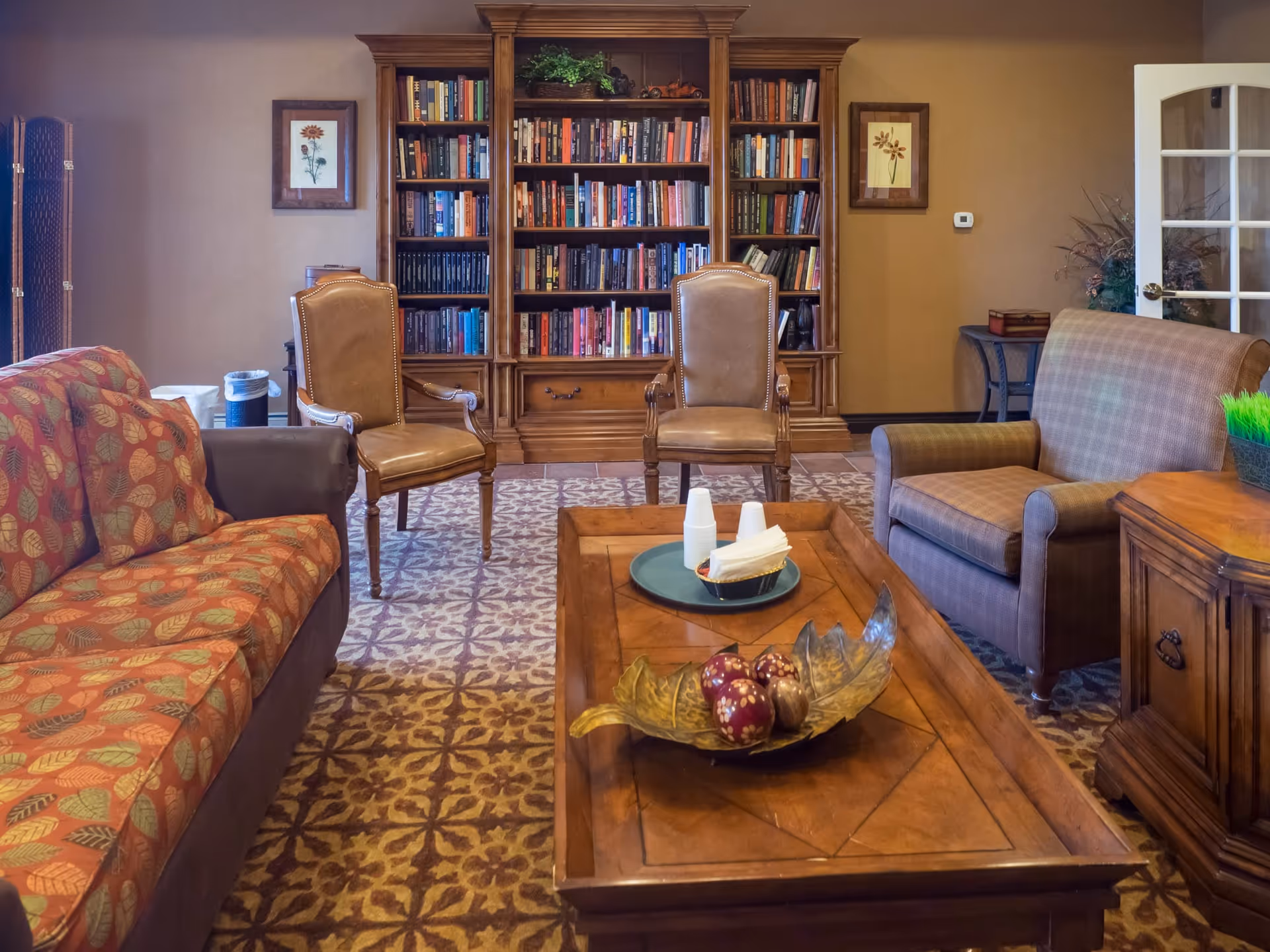 A cozy living room with a patterned carpet, a wooden coffee table with decorative items, a red and brown patterned sofa, two leather armchairs, a plaid armchair, and a large wooden bookshelf filled with books against the wall. There are framed floral pictures on the wall and a glass-paneled door on the right side.