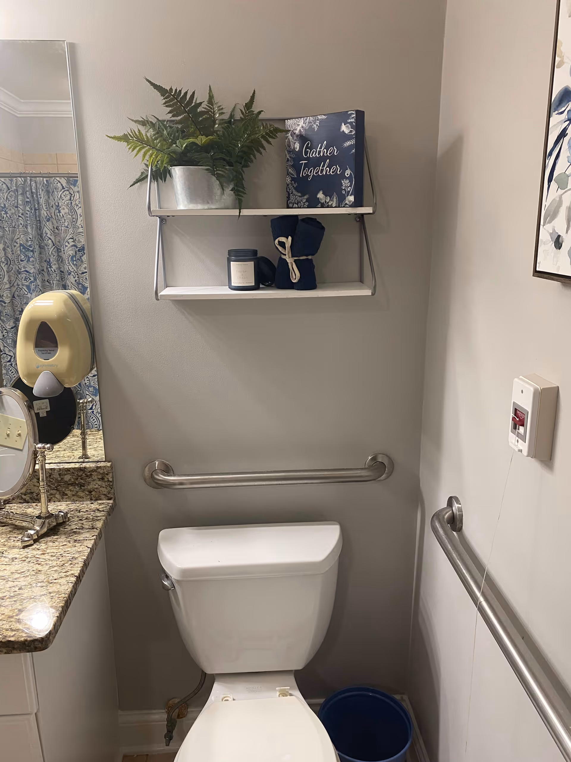 A small bathroom with a white toilet, grab bars on both sides, a granite countertop with a mirror and soap dispenser, a blue patterned shower curtain, and a white wall shelf holding a potted plant, a candle, two rolled towels, and a decorative sign that says 'Gather Together'.