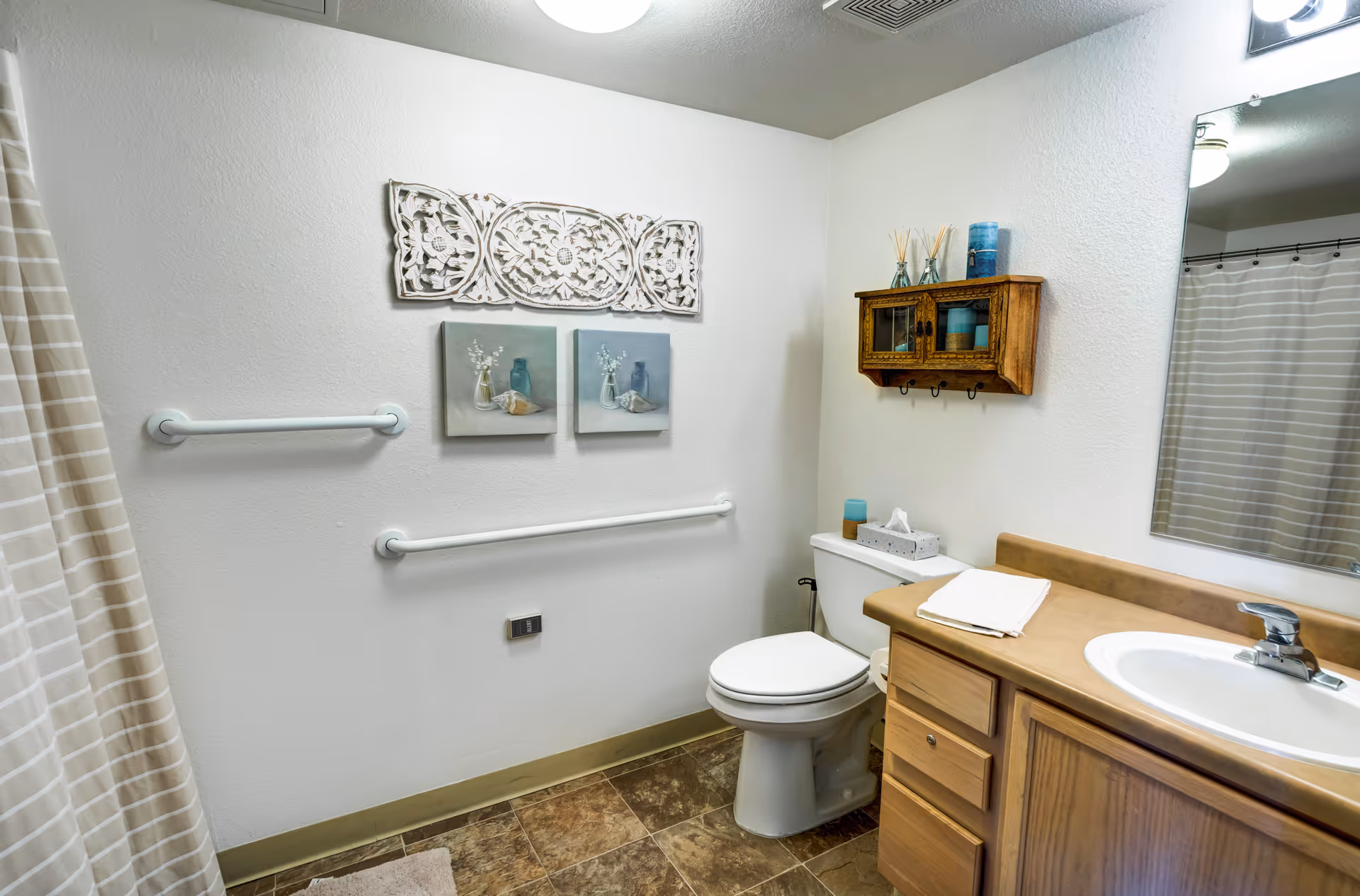 A clean and well-lit bathroom with a toilet, a sink with a wooden cabinet, a mirror, and a shower with a striped curtain. The walls are decorated with two small paintings of vases and flowers, a carved wooden wall art piece, and a small wooden cabinet holding candles and reed diffusers. There are two white grab bars on the wall near the toilet.