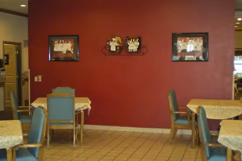 Interior view of a dining area with several tables covered with light-colored tablecloths and blue cushioned chairs. The walls are painted dark red and decorated with framed pictures and a small decorative piece. The floor is tiled, and there is a doorway visible on the left side.