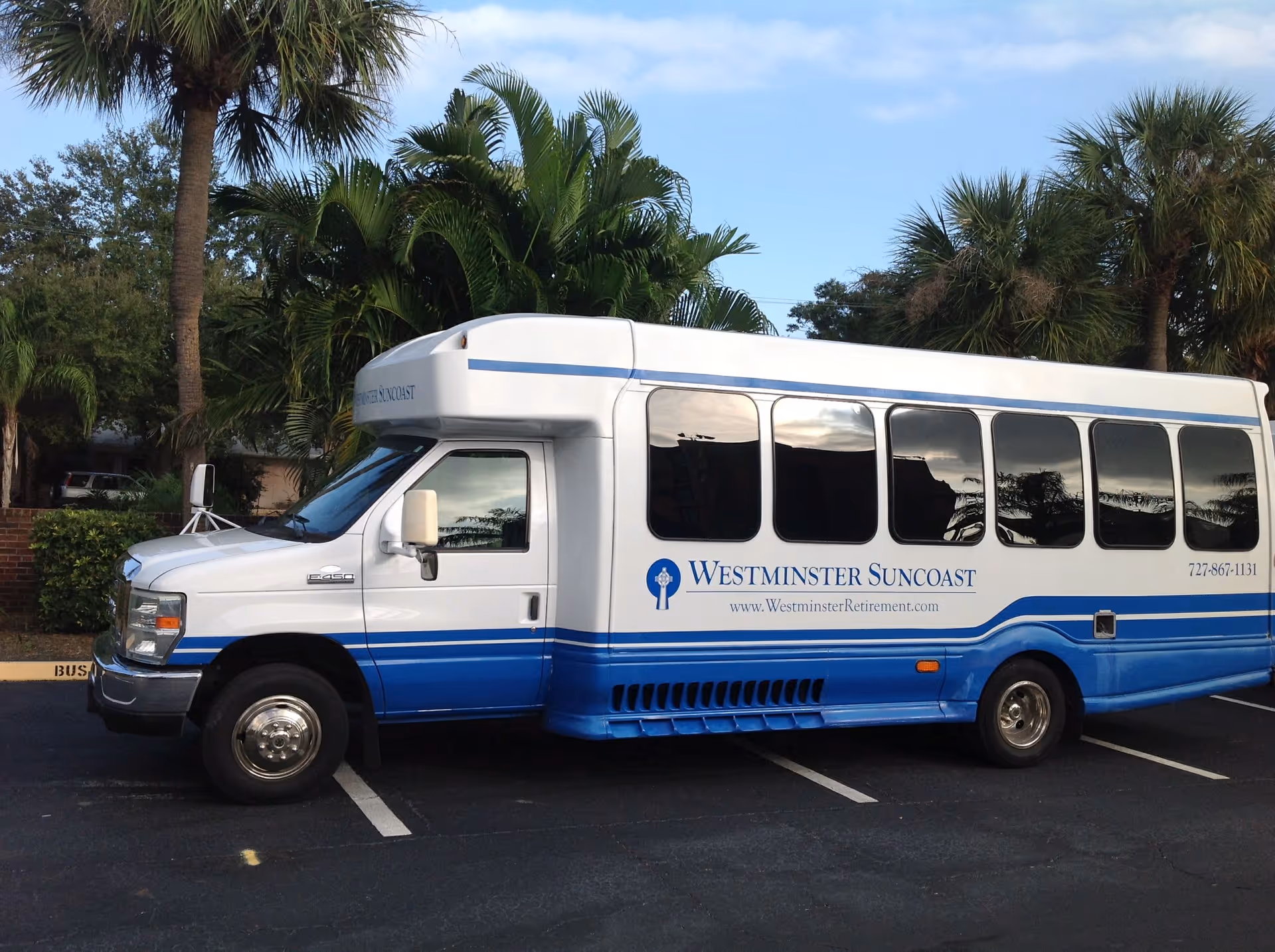 A white and blue shuttle bus parked in a lot with palm trees and greenery in the background. The bus has the text 'Westminster Suncoast' along with the website 'www.WestminsterRetirement.com' and a phone number '727-867-1131' on its side.