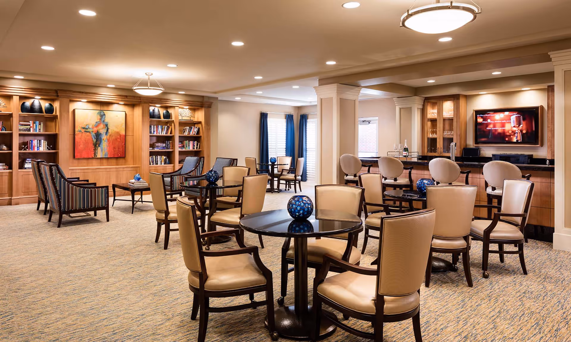 Spacious, well-lit communal lounge with round tables and upholstered chairs, a bar area, bookshelves and decorative artwork.