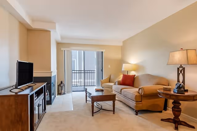 A cozy living room with a beige sofa adorned with a red pillow, a wooden coffee table, and a round side table with a lamp and a vintage rotary phone. A flat-screen TV sits on a wooden cabinet opposite the sofa. The room has beige walls and carpet, with a sliding glass door leading to a balcony.