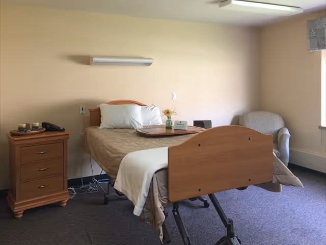 A simple senior living bedroom with a hospital-style adjustable bed covered with beige bedding and white pillows. A wooden nightstand with drawers is beside the bed, holding a phone and some candles. There is a small vase with flowers and a tray on the bed. A cushioned armchair is placed near a window with a patterned valance, and the room has beige walls and carpeted floor.