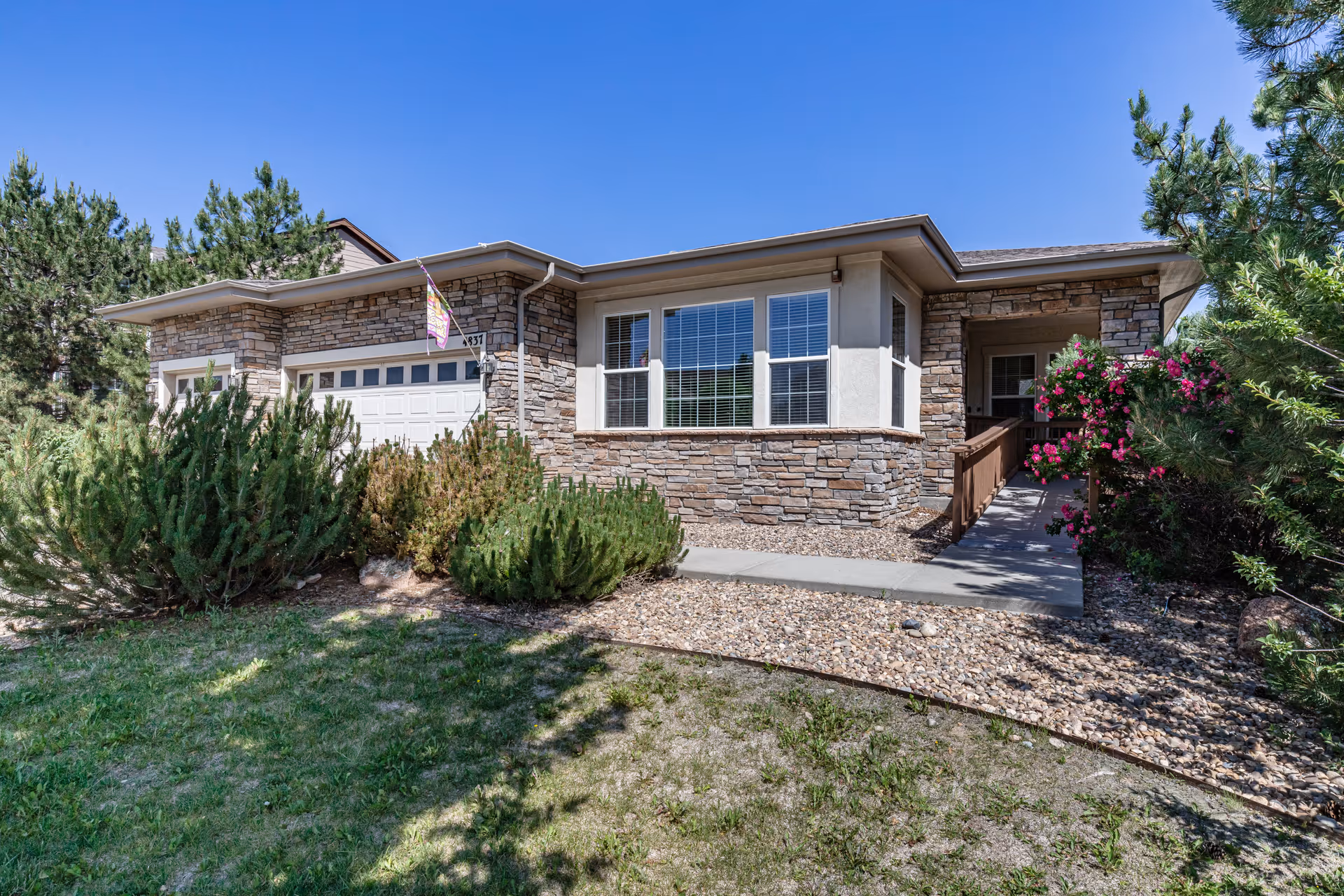 Exterior view of a single-story senior living facility building with stone and stucco facade, a white garage door, large windows, a ramp leading to the entrance, surrounded by green shrubs and flowering plants under a clear blue sky.