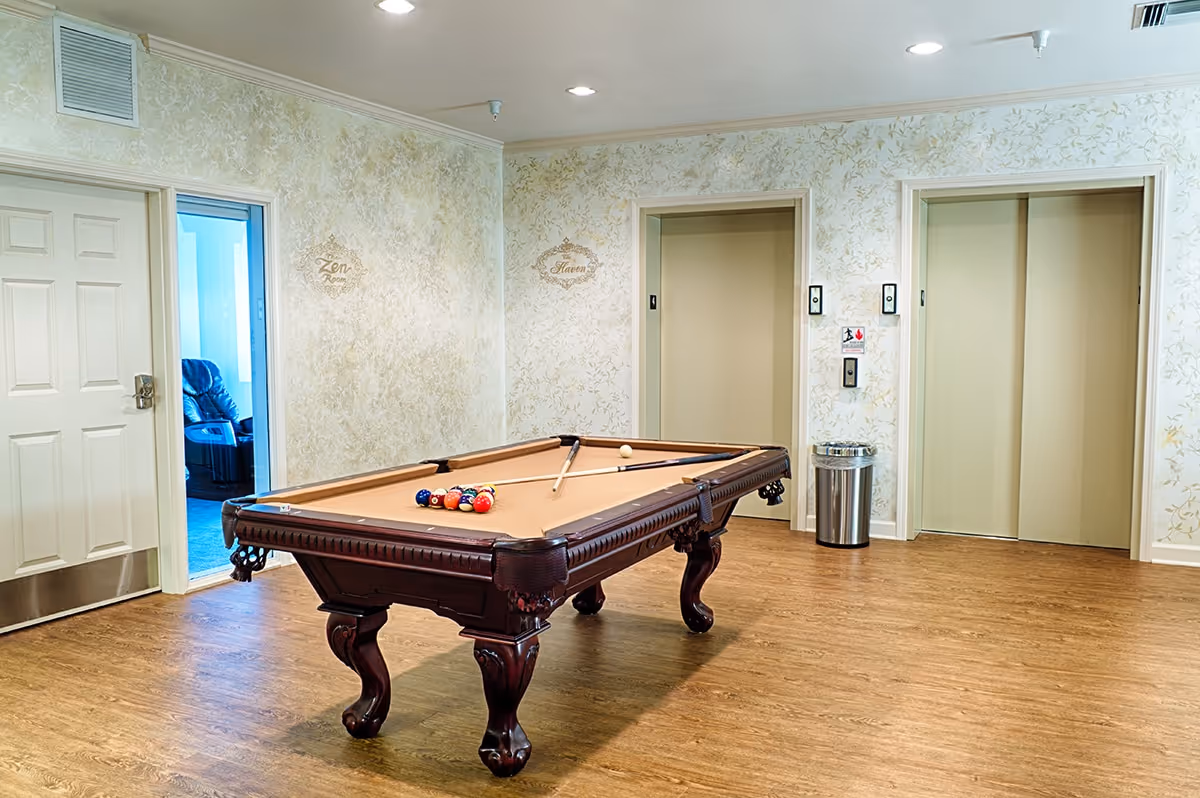 A pool table sits in a well-lit common room with patterned wallpaper, elevator doors, and a doorway to an adjacent seating area.